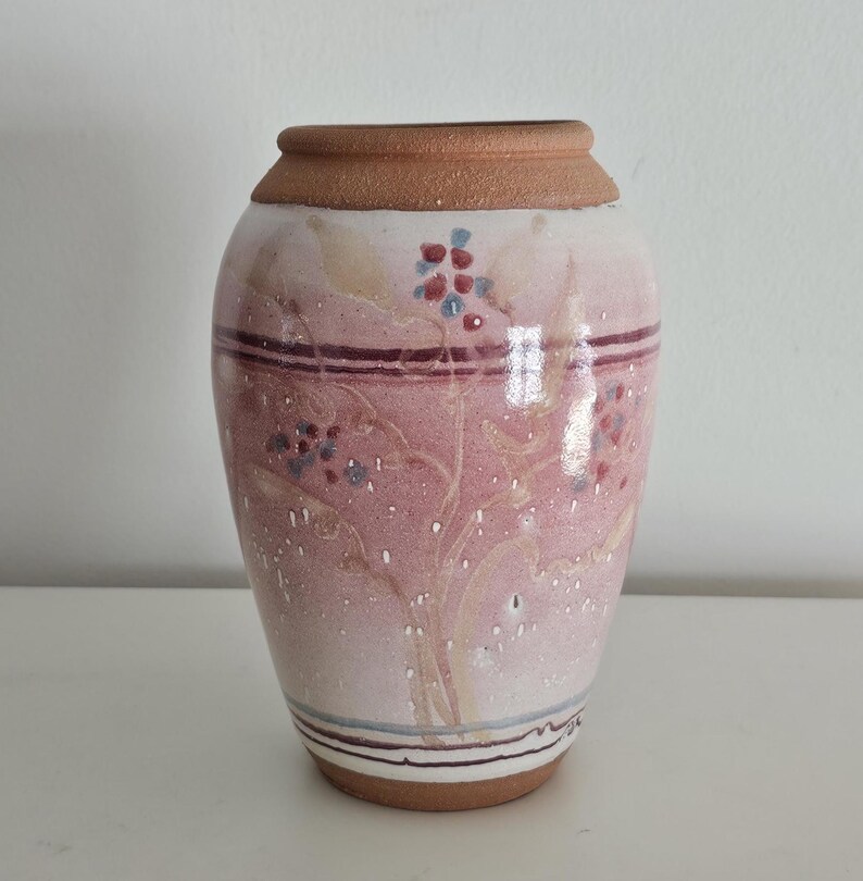 May include: A ceramic vase with a wide base and a narrow neck. The vase is painted with a pink and white floral design, with a brown rim and base. The vase is approximately 7 inches tall.