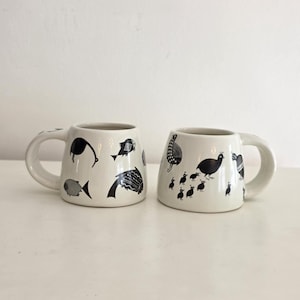 May include: Two off-white ceramic mugs with a wide base and a thick handle. The left mug features black graphic illustrations of various fish. The right mug displays black graphic illustrations of a bird and several smaller chicks in a line.