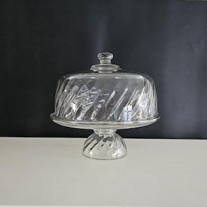 May include: Clear glass cake stand with a domed lid. The stand features a spiral design on the base, the plate, and the lid. The lid has a small knob on top. The cake stand is on a white surface with a black background.