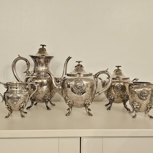 May include: A silver tea set with a teapot, creamer, sugar bowl, and a matching serving tray. The set is decorated with an embossed floral pattern.