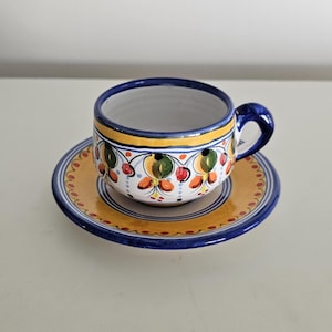 May include: A hand-painted ceramic teacup and saucer set. The cup and saucer feature a white base with a yellow border and a blue rim. The cup and saucer are decorated with colorful floral patterns in green, red, and orange.