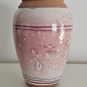 May include: A handcrafted ceramic vase with a reddish-pink glaze and speckled white accents. The vase features horizontal bands of dark red and light blue near the top and bottom, and a brown rim and base. The vase is a decorative home decor item.
