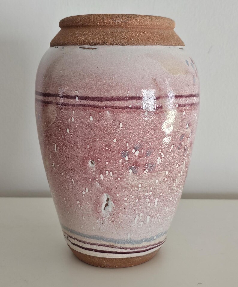May include: A handcrafted ceramic vase with a reddish-pink glaze and speckled white accents. The vase features horizontal bands of dark red and light blue near the top and bottom, and a brown rim and base. The vase is a decorative home decor item.