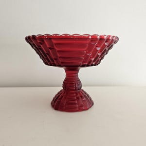 May include: A ruby red, vintage glass pedestal bowl. The bowl has a diamond pattern and a scalloped rim. The base is a detailed, tiered design. The bowl is likely used for serving fruit or displaying decorative items.