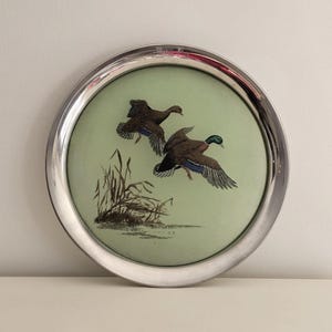 May include: A silver-plated round tray with a green background and a painted image of two ducks in flight. The ducks are brown and green with blue wings. The tray has a raised rim.
