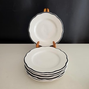 May include: A set of white ceramic plates featuring a scalloped edge design with a thin, dark navy blue rim. The plates are stacked in a pile, with one plate positioned upright on a wooden stand to display the circular shape and edge detail.