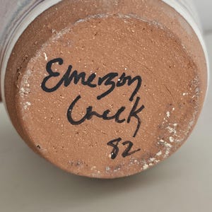 May include: Close-up of the bottom of a ceramic mug. The base is a reddish-brown color with the handwritten text "Emerson Creek" and the number "82" in black ink. The mug's exterior has a white and light-colored design.