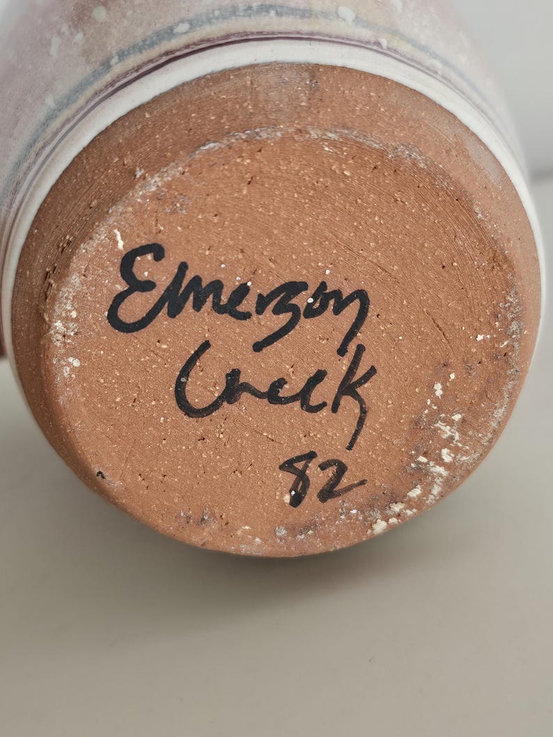 May include: Close-up of the bottom of a ceramic mug. The base is a reddish-brown color with the handwritten text "Emerson Creek" and the number "82" in black ink. The mug's exterior has a white and light-colored design.