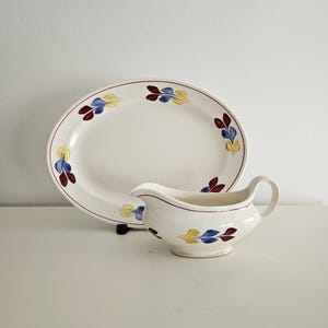 May include: A white oval serving platter with a red, yellow, and blue leaf design around the rim. A white gravy boat with the same leaf design is in front of the platter.