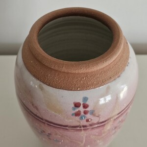May include: A handmade ceramic vase with a wide opening and a rounded body. The vase features a light pink and white base with a brown rim. Delicate floral patterns in red and blue adorn the surface, adding a touch of artistry to this decorative piece.
