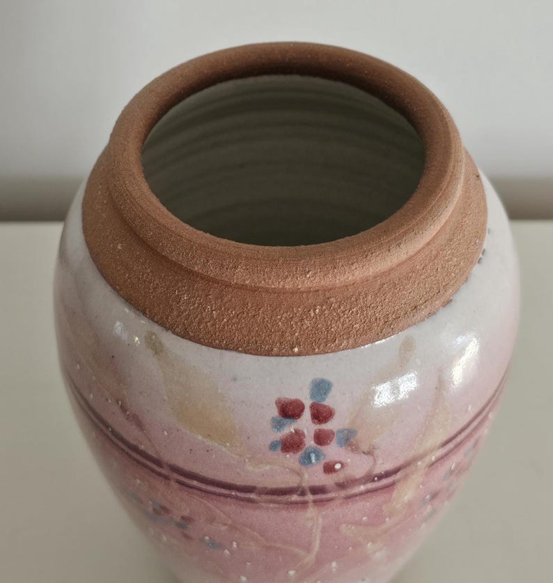 May include: A handmade ceramic vase with a wide opening and a rounded body. The vase features a light pink and white base with a brown rim. Delicate floral patterns in red and blue adorn the surface, adding a touch of artistry to this decorative piece.