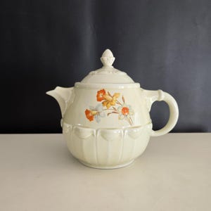 May include: Cream-colored ceramic teapot with a lid and handle. The teapot features a floral design with orange, yellow, and white flowers. The body of the teapot has a scalloped design. The lid is topped with a decorative finial.