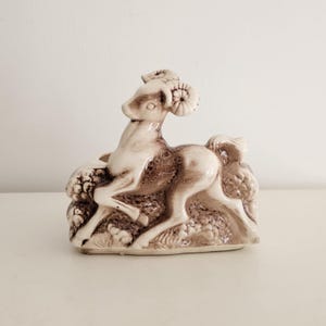 May include: A ceramic ram figurine in shades of brown and cream. The ram is depicted in a dynamic pose, with its head turned and horns curled. The figurine is set on a base with textured details, suggesting a natural setting.