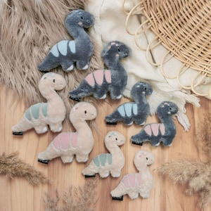 Könnte beinhalten: Eine Sammlung von Plüsch-Dinosaurierspielzeugen in verschiedenen Farben, darunter Grau, Rosa und Beige. Die Dinosaurier haben farblich abgesetzte Akzente auf dem Rücken und sind auf einer Holzoberfläche angeordnet. Einige haben blaue, rosa oder grüne Akzente.
