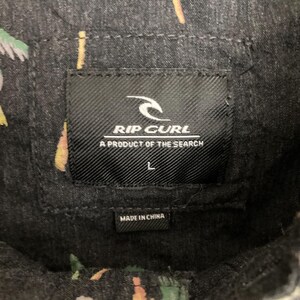 May include: Close-up of a black Rip Curl shirt label. The label features the Rip Curl logo and the text "A PRODUCT OF THE SEARCH". The size "L" is printed on the label. The shirt is made in China.