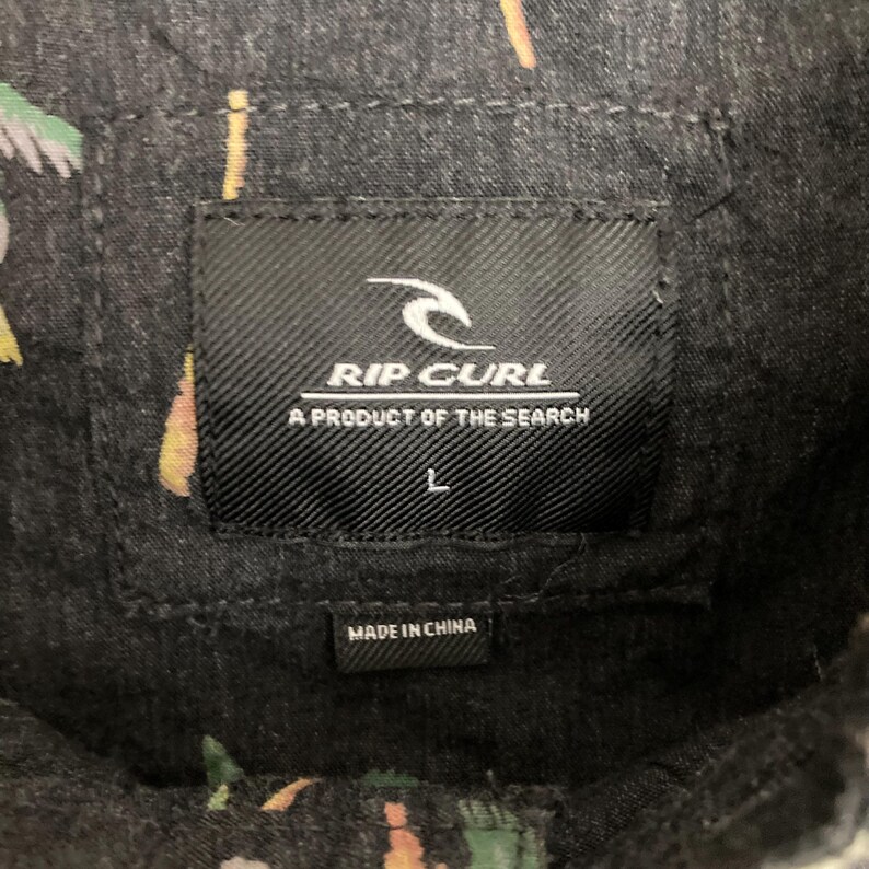 May include: Close-up of a black Rip Curl shirt label. The label features the Rip Curl logo and the text "A PRODUCT OF THE SEARCH". The size "L" is printed on the label. The shirt is made in China.