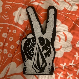 May include: Black and silver peace sign hand with the word "PEACE" and a logo. The hand is on a patterned orange and white background. The peace sign is a foam hand.