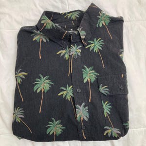 May include: A dark gray button-down shirt with a tropical palm tree pattern. The shirt features a classic collar, a chest pocket, and black buttons. The palm trees are in shades of green, yellow, and brown.