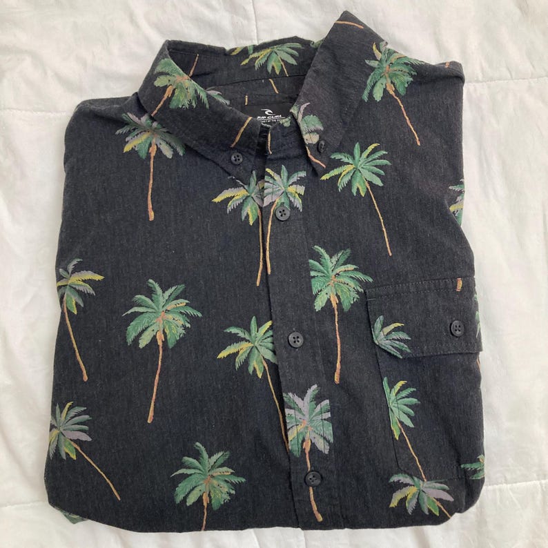 May include: A dark gray button-down shirt with a tropical palm tree pattern. The shirt features a classic collar, a chest pocket, and black buttons. The palm trees are in shades of green, yellow, and brown.