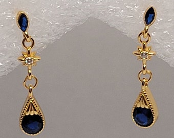 May include: A pair of gold-colored drop earrings. Each earring features a teardrop-shaped, dark blue gemstone at the bottom, a star-shaped accent with a small clear stone, and a small, dark blue marquise-shaped stone at the top.