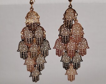 Peut inclure: Boucles d'oreilles pendantes dorées avec un motif en cascade de mains de Hamsa ajourées. Les boucles d'oreilles présentent un motif filigrané détaillé et sont suspendues à des crochets d'oreilles dorés. Elles sont présentées sur un fond neutre.