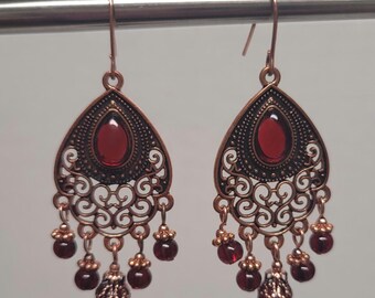 May include: A pair of dangle earrings with a filigree design and red gemstone accents. The earrings are made of copper and have a dark brown finish.