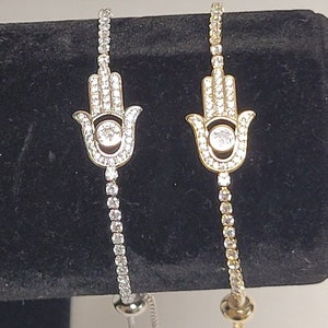 May include: Two adjustable bracelets with a hamsa hand charm. One bracelet is silver with clear crystals and the other is gold with clear crystals.
