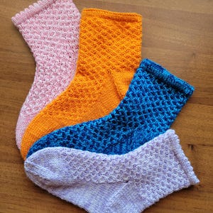 May include: Four pairs of knitted socks in different colours: pink, orange, blue, and purple. The socks have a textured pattern and are folded in a fan shape.