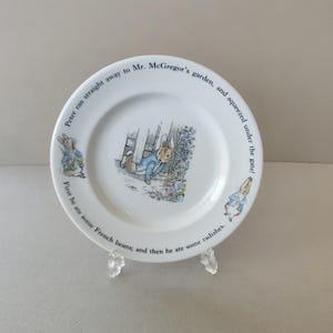 May include: White ceramic plate with illustrations of Peter Rabbit and text from the children's story. The plate features Peter Rabbit squeezing under a gate, with additional illustrations and text around the rim.