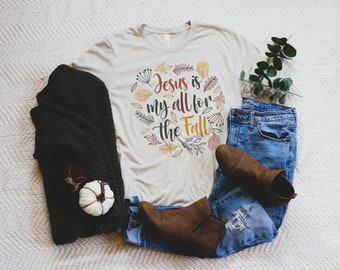 Puede incluir: Una camiseta beige con un diseño gráfico que dice "Jesus is my all for the Fall" rodeada de hojas de otoño en tonos de rojo, naranja, amarillo y verde.