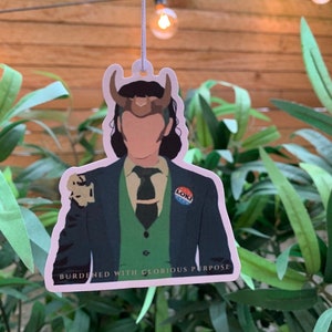 May include: A white car air freshener featuring a cartoon illustration of Loki from the Marvel Cinematic Universe. He is wearing a green suit, a green vest, and a gold crown. The text "BURDENED WITH GLORIOUS PURPOSE" is printed below the image. The text "LOKI" is printed on a blue button on his suit.