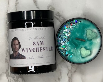 May include: A black candle jar with a label that reads "The Marvelous Candle Studio" and "Smells Like Sam Winchester" with a picture of the character Sam Winchester from the TV show Supernatural. The candle is soy wax, vegan, cruelty-free, and hand-poured in London. The candle is amber, torfa, and cedar scented. The candle is next to a teal green candle with glitter and two heart-shaped wax decorations.