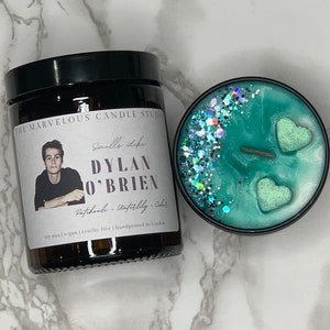 May include: A soy wax candle in a glass jar with a black lid. The candle is decorated with glitter and two green heart-shaped embellishments. The label on the jar reads "The Marvelous Candle Studio" and "Smells Like Dylan O'Brien Patchouli - Waterlily - Cedar".