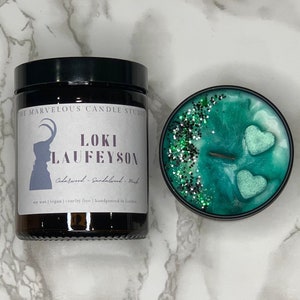 May include: A green and black candle with a glitter design and two heart-shaped accents. The candle jar has a label that reads "The Marvelous Candle Studio" and "Loki Laufeyson Cedarwood - Sandalwood - Musk" with a silhouette of a horned figure. The label also states "soy wax | vegan | cruelty free | handpoured in London."