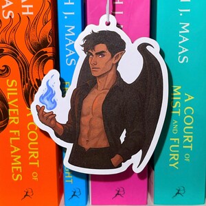 May include: A bookmark featuring a male character with dark hair, wings, and a blue flame, set against the backdrop of colorful books by Sarah J. Maas. The books are in orange, blue, and pink.