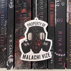 May include: A white bookmark featuring a gas mask design and the text "PROPERTY OF MALACHI VIZE" hangs in front of a row of books. The gas mask is black with red splatter details. The books have dark covers.