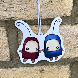 May include: A white car air freshener with two cartoon characters. The characters are holding hands and have big eyes. One character is wearing a red suit and the other is wearing a blue suit.