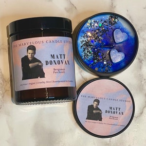 May include: Two soy wax candles with a black and white photo of a person on the front. The candle on the left is in a clear glass jar with a black lid. The candle on the right is in a black jar with a blue and white design and glitter. Both candles have the text "The Marvelous Candle Studio" and "Matt Donovan" on the front. The text "Bergamot Patchouli" is also on the front of both candles. The text "Soy wax | Vegan | Cruelty free | Handpoured in London" is on the front of the candle on the left. The text "Soy wax | Vegan | Cruelty free | Handpoured in London" is on the front of the candle on the right.