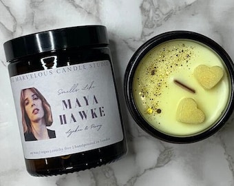 May include: A black glass candle jar with a white label that reads "Smells Life MAYA HAWKE Lychee & Peony" and "Soy wax | Vegan | Cruelty free | Handpoured in London". The candle is topped with a yellow wax and two heart-shaped wax decorations.