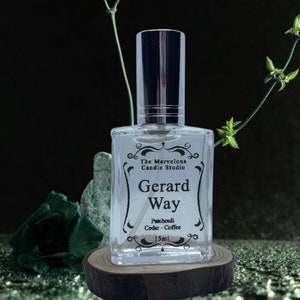 May include: A clear glass bottle with a silver spray nozzle and a black label that reads "The Marvelous Candle Studio Gerard Way Patchouli Cedar - Coffee 15ml". The bottle is sitting on a wooden round slice.
