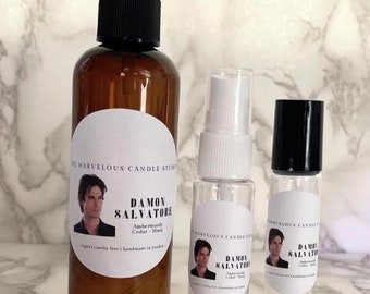 May include: Three glass bottles with labels featuring a man's face and the text "Damon Salvatore" with the words "Amberwoods Cedar - Musk" and "vegan | cruelty free | handmade in London". The bottles are different sizes and have black caps. The bottles are on a white marble surface.
