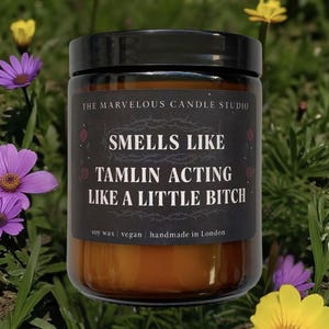 Puede incluir: Vela de cera de soja en un tarro de vidrio transparente con tapa negra. La etiqueta dice "SMELLS LIKE TAMLIN ACTING LIKE A LITTLE BITCH". Hecha a mano en Londres, rodeada de flores.