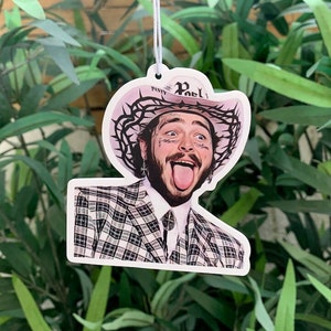 May include: A car air freshener featuring a portrait of a man with tattoos and a pink cowboy hat with the word "Pristy" on it. The man is wearing a black and white plaid suit and has his tongue sticking out.