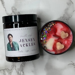 May include: A soy wax candle with a black lid and a label that reads "The Marvelous Candle Studio" and "Smells Like Jensen Ackles". The candle is decorated with glitter and two gold hearts. The label also states "Amber - Coffee - Patchouli" and "Soy wax | Vegan | Cruelty free | Handpoured in London."