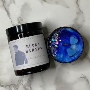 May include: A brown glass candle jar with a black lid. The label reads "The Marvelous Candle Studio" and "Bucky Barnes" in white text. The candle is soy wax, vegan, cruelty-free, and hand-poured in London. The candle is blue with silver glitter and two blue heart-shaped decorations.