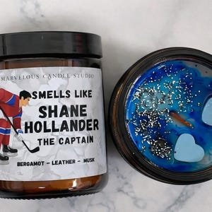 Puede incluir: Dos velas perfumadas. Una está en un tarro de cristal con tapa negra y una etiqueta que dice "Smells Like Shane Hollander The Captain". La otra vela está en un recipiente negro con cera azul y detalles en forma de corazón.