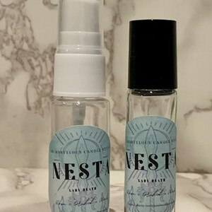 Puede incluir: Dos botellas de vidrio transparente con tapas negras. Las botellas tienen etiquetas con el texto "The Many Eloquent Candles by Nesta Lady of the Obsidian Flame" y "Nesta Lady of the Obsidian Flame".