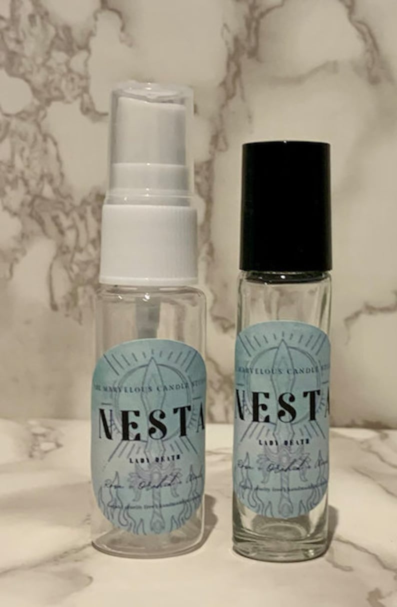 Puede incluir: Dos botellas de vidrio transparente con tapas negras. Las botellas tienen etiquetas con el texto "The Many Eloquent Candles by Nesta Lady of the Obsidian Flame" y "Nesta Lady of the Obsidian Flame".