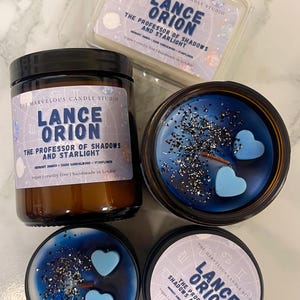 Op de afbeelding: Een assortiment geurkaarsen en wax melts van The Marvelous Candle Studio. De producten zijn voorzien van de tekst "Lance Orion: The Professor of Shadows and Starlight". De kaarsen zijn blauw met hartvormige wax melts en glitter.