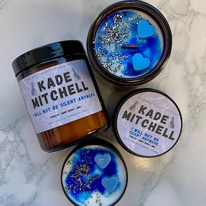 May include: An assortment of scented candles in glass jars and tins. The candles feature blue wax with heart-shaped accents and silver glitter. The labels read "Kade Mitchell I Will Not Be Silent Anymore".